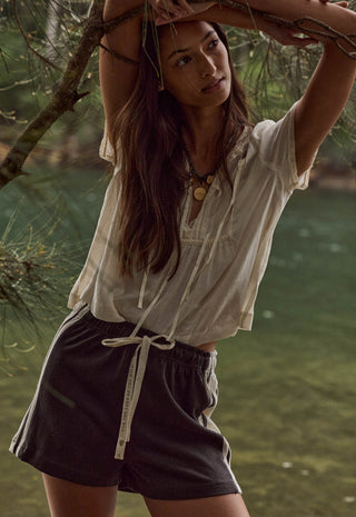 a-woman-outdoors-near-water-and-tree-branches-wearing-a-loose-white-short-sleeve-top-and-dark-charcoal-drawstring-shorts