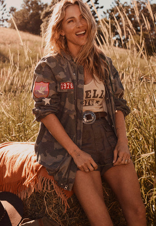 a-woman-sitting-outdoors-in-a-grassy-field-wearing-a-camouflage-utility-jacket-over-a-beige-graphic-tank-top-and-dark-shorts-with-a-bright-orange-fringed-blanket-beside-her