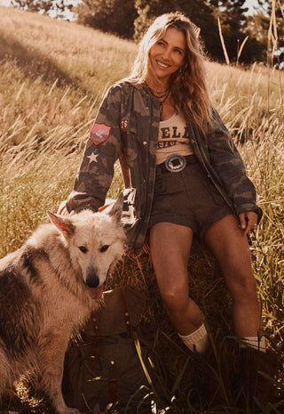 a-woman-sitting-in-a-grassy-field-with-a-dog-wearing-a-camouflage-bomber-jacket-over-a-graphic-tee-and-shorts